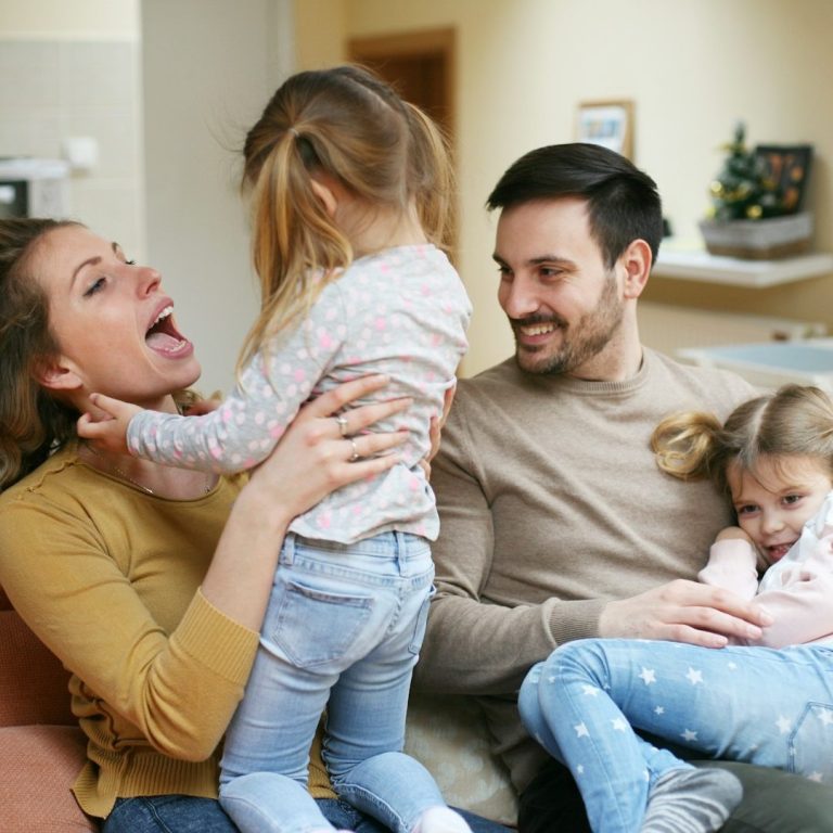 Eine Familie spielt lachend auf der Couch mit zwei kleinen Mädchen.