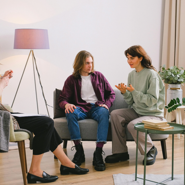 Eine Mann und eine Frau sprechen, eine weitere Frau schreibt Notizen. Es handelt sich um ein Therapiesetting.