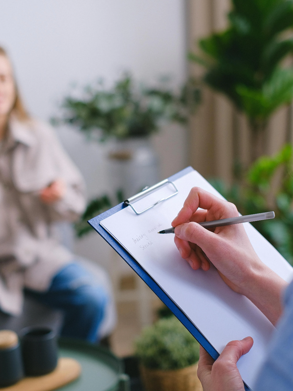 Eine Hand hält einen Stift über ein Notizblatt, während im Hintergrund eine Person sitzt.