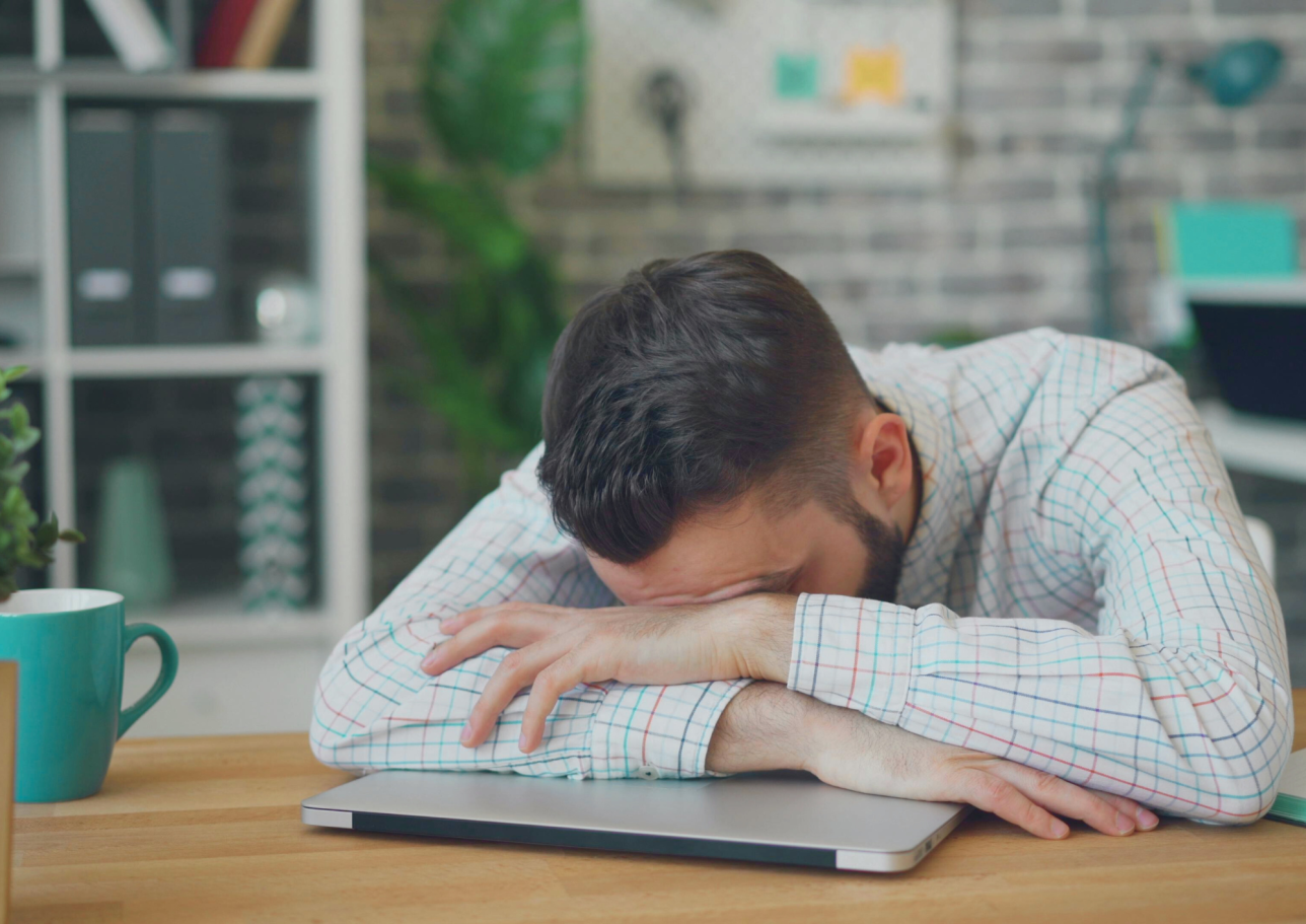 Ein Mann mit Bart lehnt müde auf einem Laptop, umgeben von Büromaterialien.