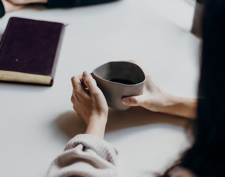 Eine Person hält eine graue Tasse in den Händen, während ein Buch daneben liegt.