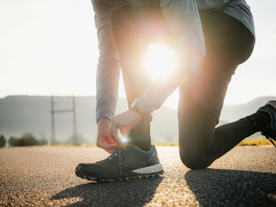 Person bindet die Schnürsenkel eines Sportschuhs bei Sonnenuntergang auf einer Straße.