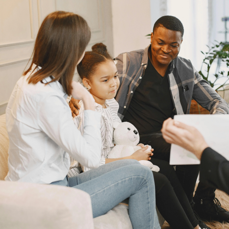 Familie sitzt zusammen, spricht und lächelt, ein Kind hält ein Stofftier.