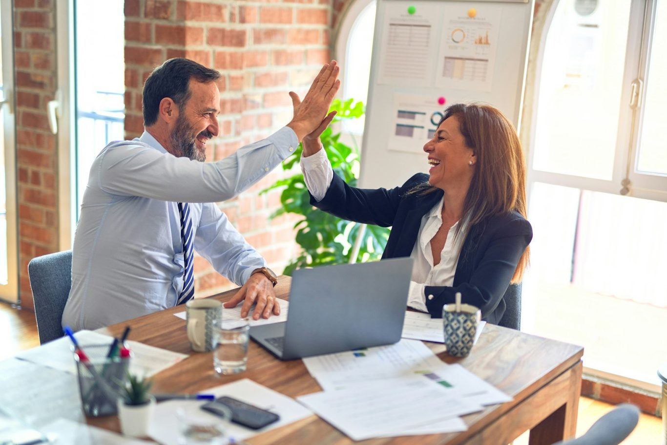 Zwei Personen im Büro geben sich einen High-Five, lächelnd am Tisch mit Laptop.